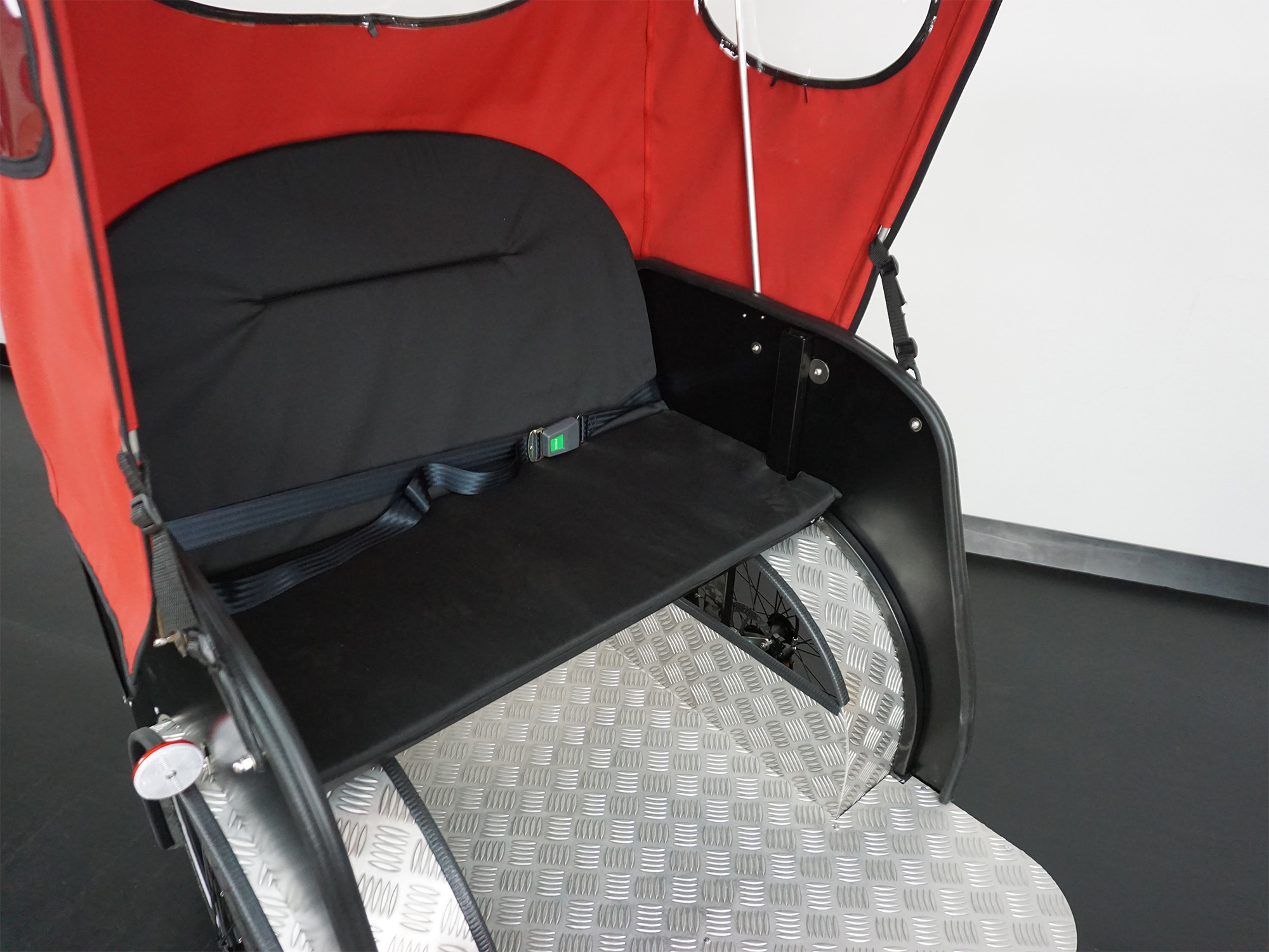 Sitzbereich eines Lastenrads mit rotem Verdeck, schwarzer Rückenlehne, Sicherheitsgurt und rutschfestem Riffelblechboden.