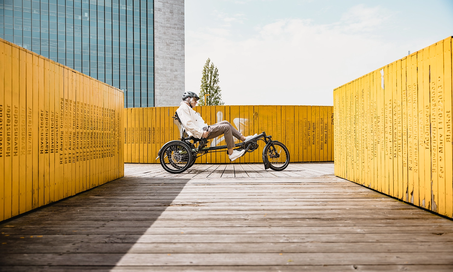 Mann fährt entspannt auf einem Liegedreirad durch eine gelbe holzinstallation in der Stadt