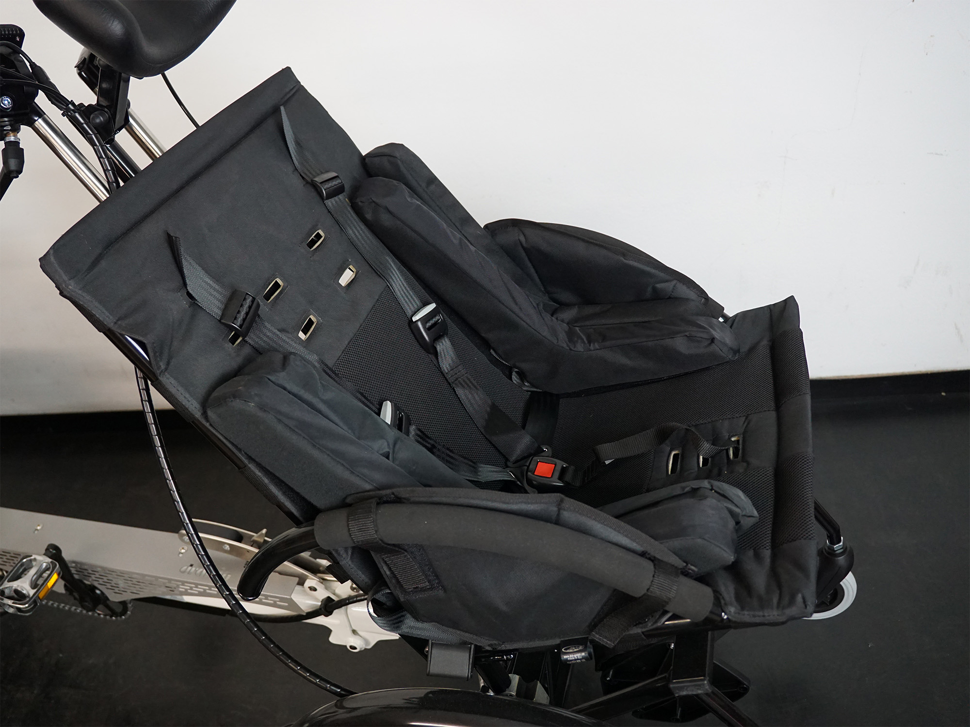 Schwarzer Spezial-Kindersitz mit Seitenhalt, Polsterung und 5-Punkt-Gurt auf einem weißen Therapierad montiert.