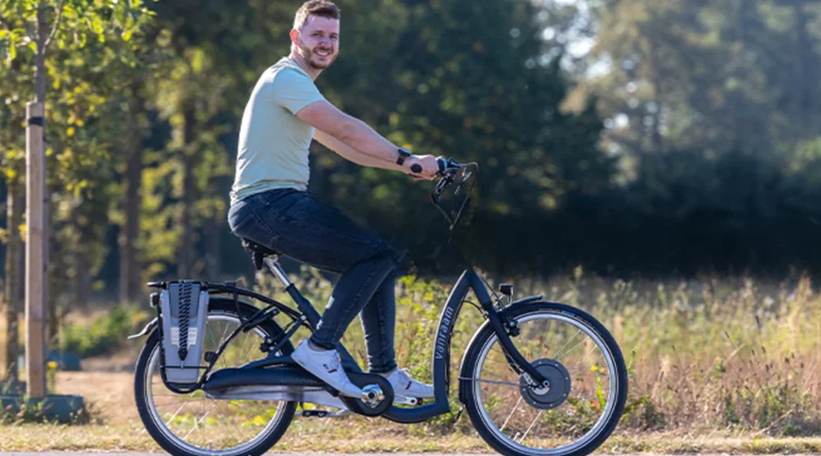 Junger Mann fährt Van Raam E-Bike auf einem Radweg durch die Natur und lächelt in die Kamera.