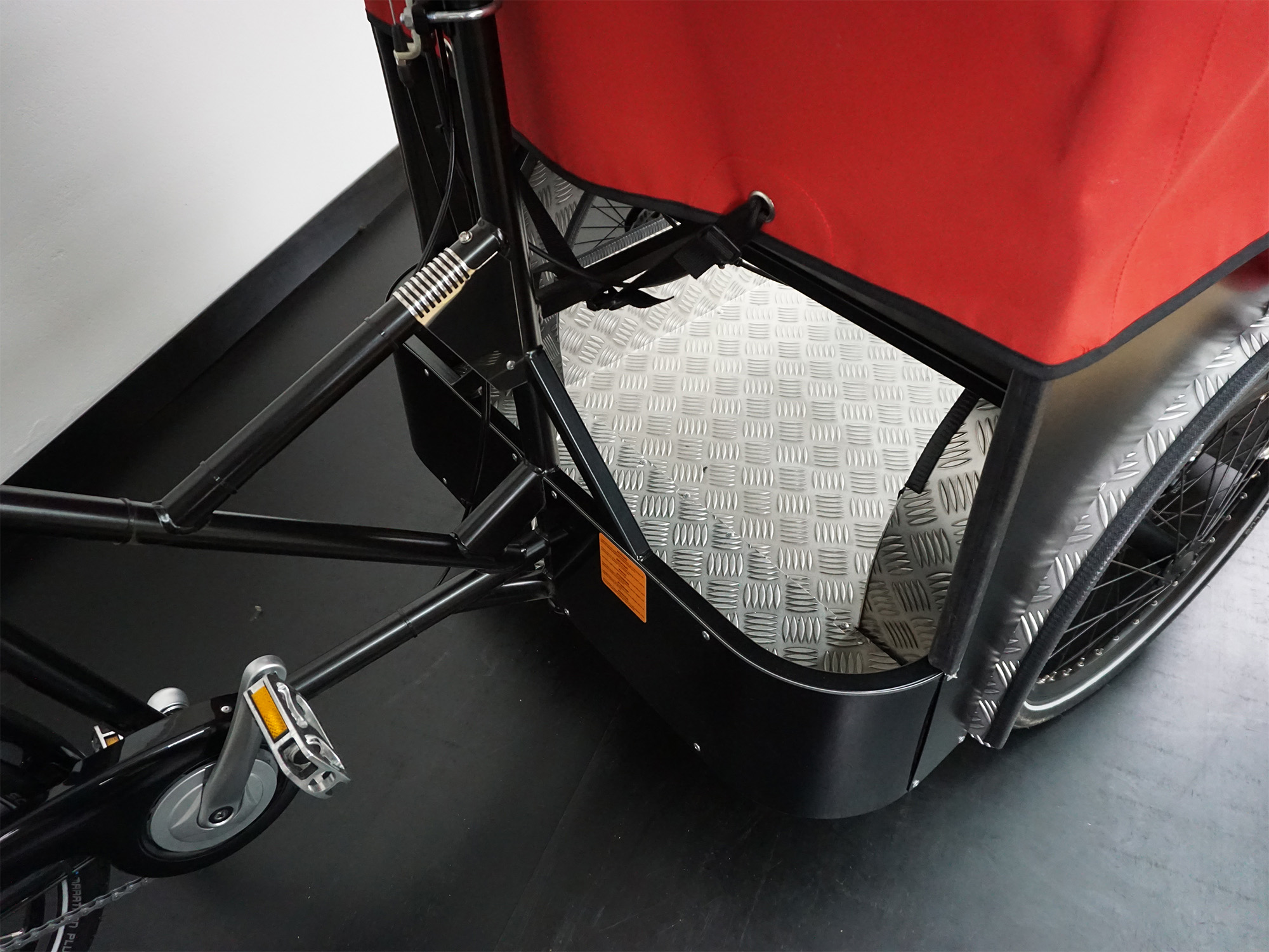 Teilansicht eines Lastenrads mit rotem Aufbau, schwarzem Rahmen, Pedal, Riffelblechboden und integriertem Transportbereich.