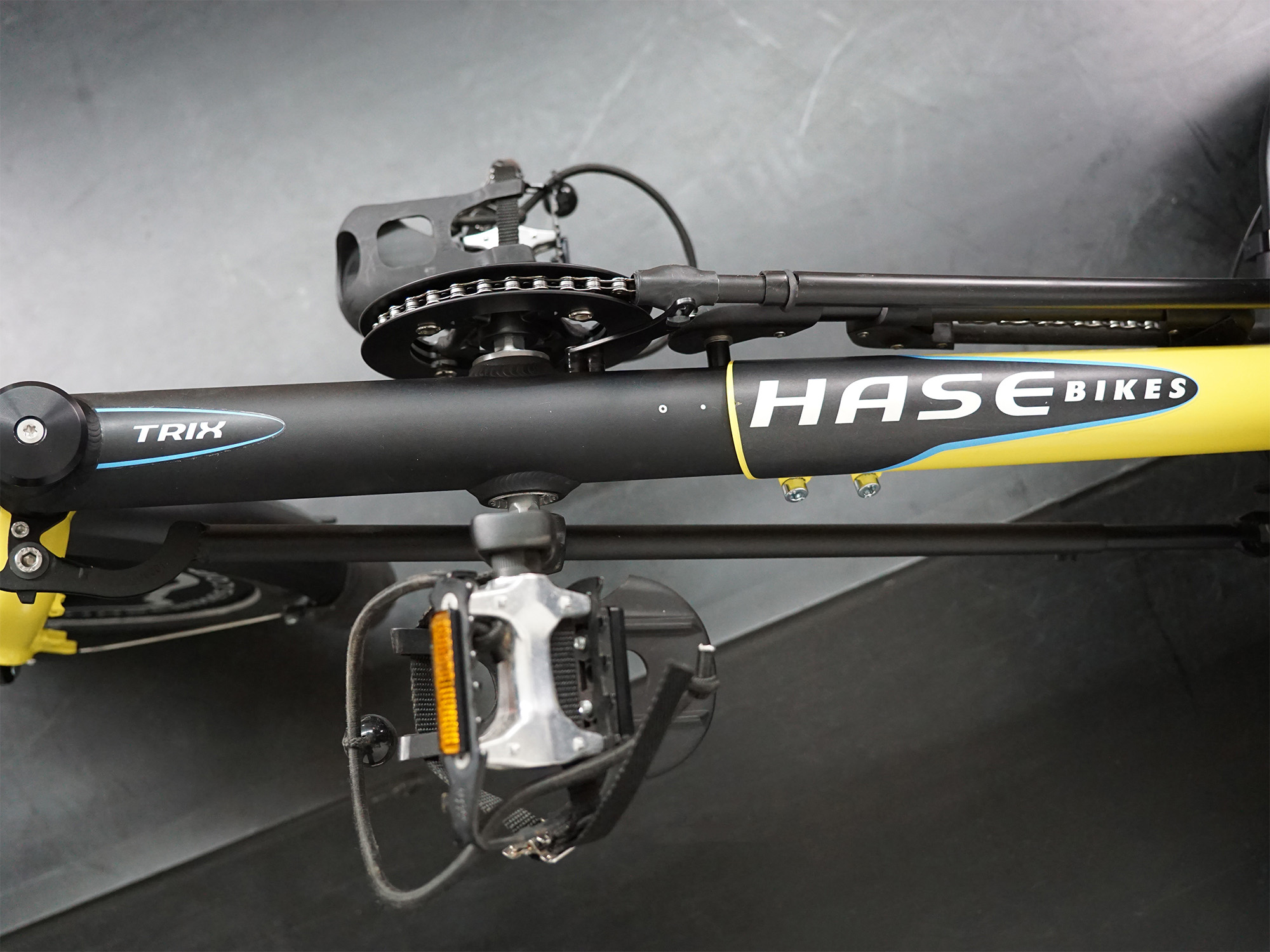 Liegerad mit schwarzem Rahmen, „HASE BIKES“- und „TRIX“-Schriftzug, Pedal mit Reflektor und Kettentrieb.