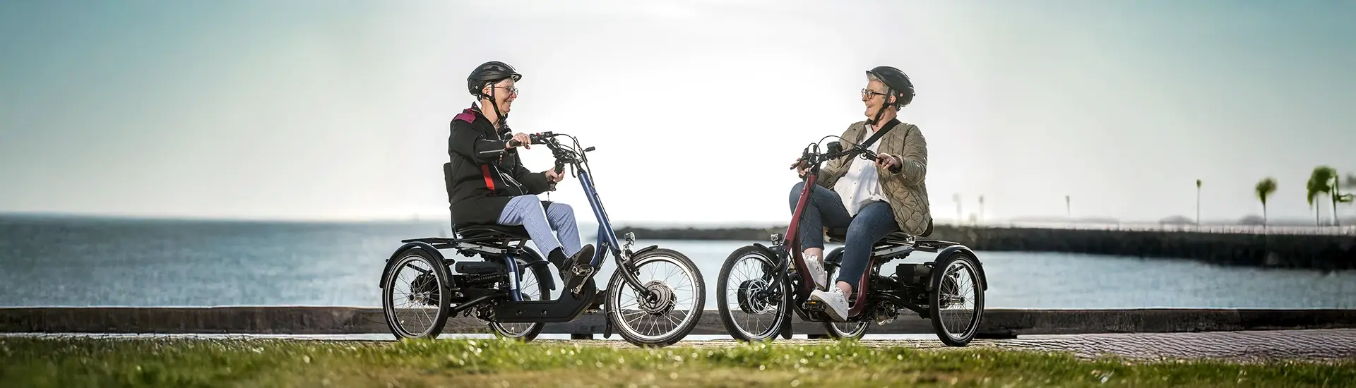 Zwei Frauen sitzen jeweils auf einem Dreirad und stehen sich gegenüber. Vor einem Gewässer