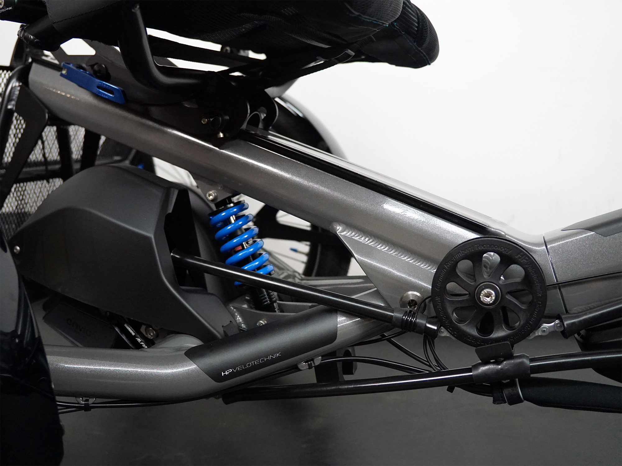 Detailansicht eines Dreirads mit blauer Federung, Zahnriemen und Kettenführung am grauen Rahmen.