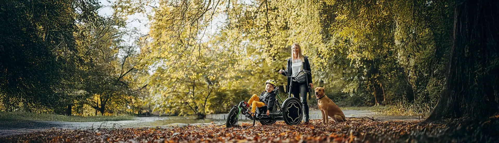 Ein Junge sitzt in einem Dreirad im Wald während die Mutter dahinter steht.