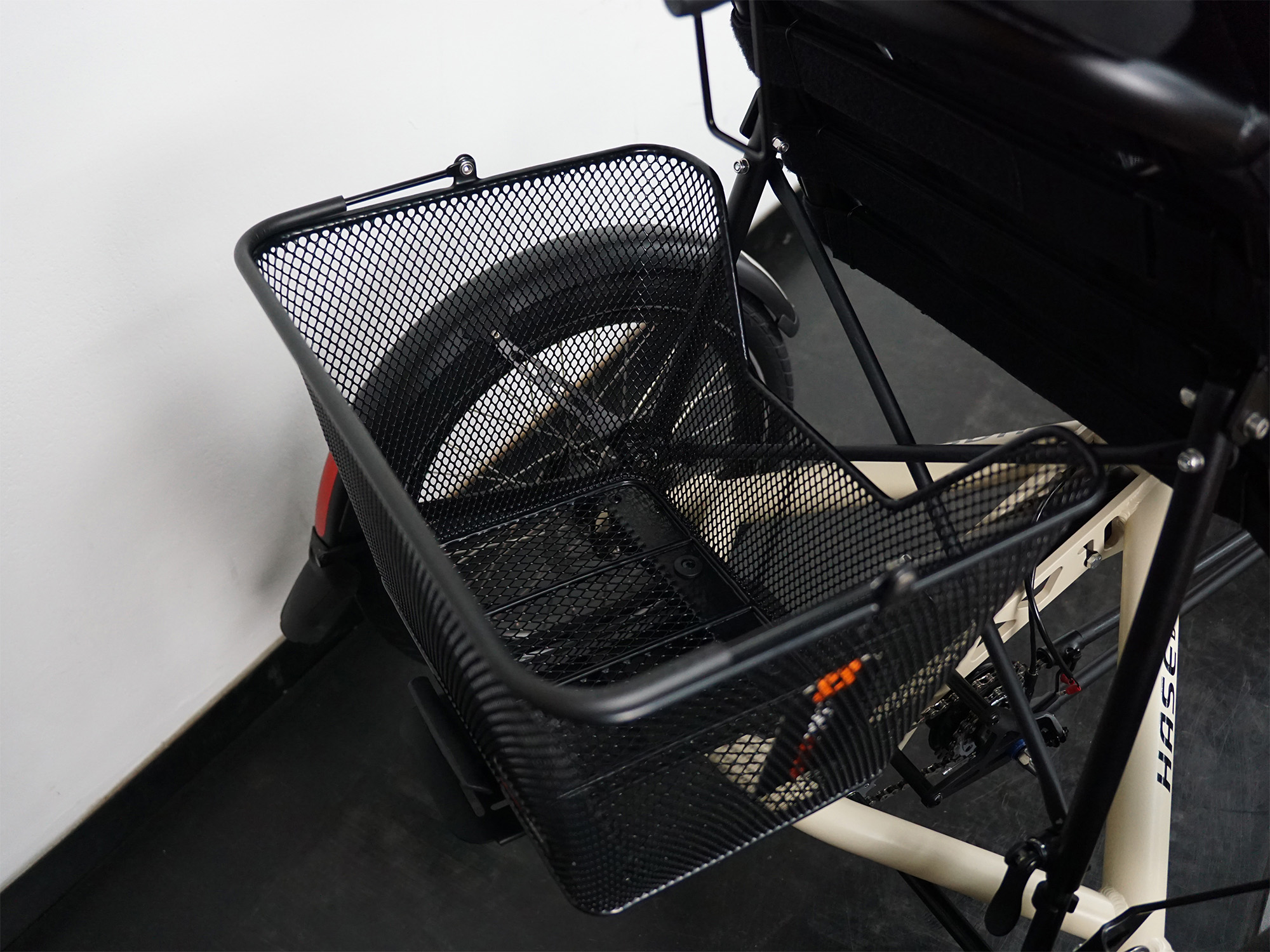 Liegedreirad mit stabilem schwarzem Gepäckkorb aus Metallgitter hinter dem Sitz, beige Rahmenfarbe, Rücklicht sichtbar.