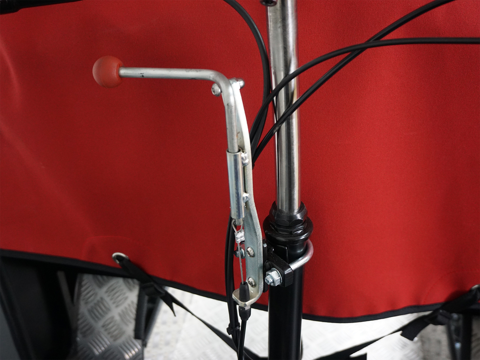 Detailaufnahme eines roten Transportrads mit mechanischer Feststellbremse, Bowdenzügen und Riffelblechboden im Hintergrund.